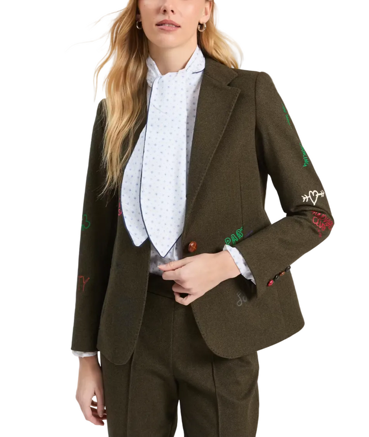A woman models an olive green tailored blazer with colorful embroidered initials on the sleeves, paired with matching trousers and a light blue patterned high-neck blouse against a plain light gray background.