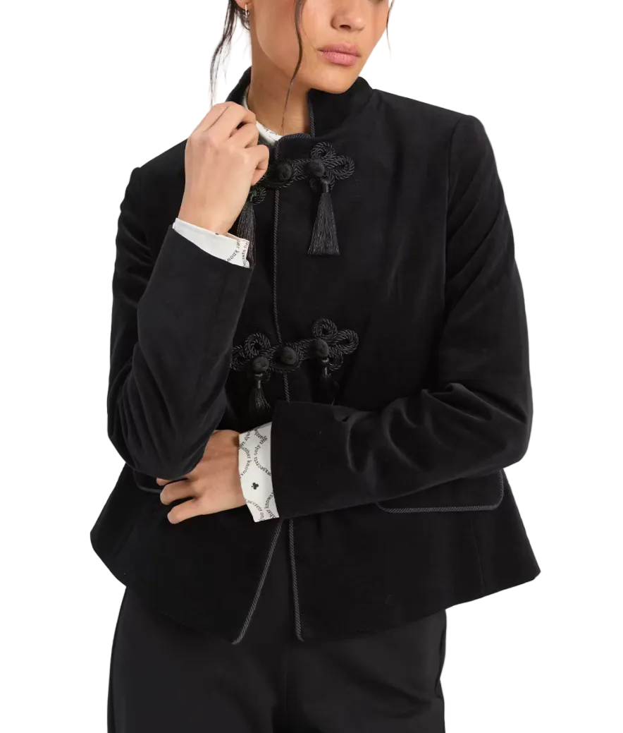 A model wearing a black velvet cropped jacket with traditional Chinese-style frog closures and tassel details, layered over a white patterned shirt.