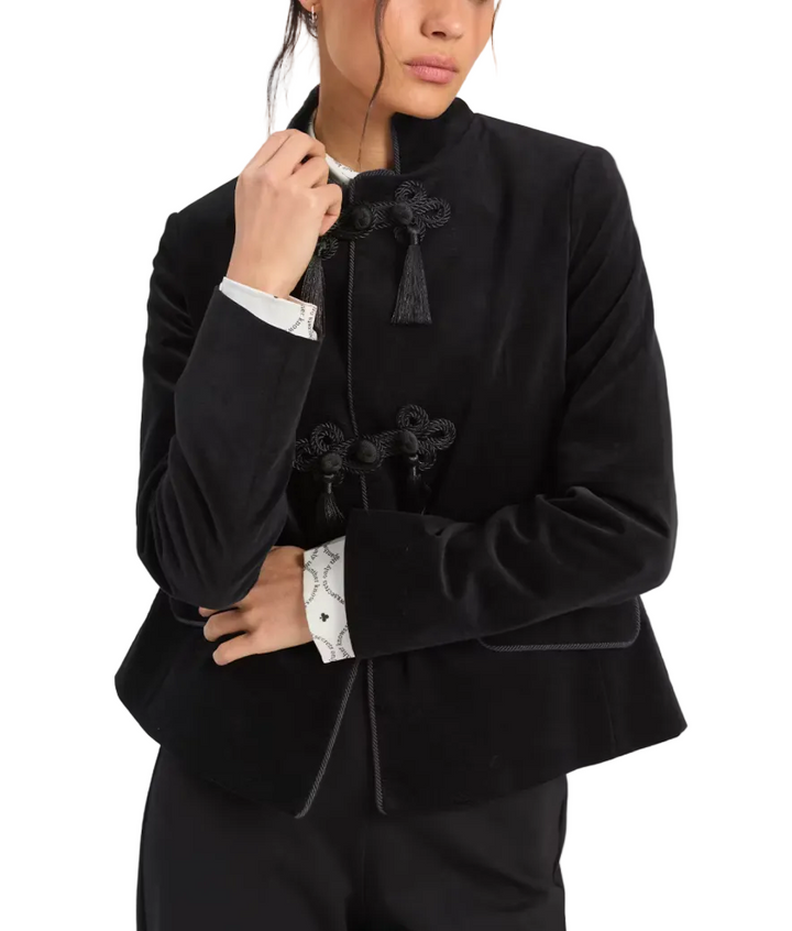 A model wearing a black velvet cropped jacket with traditional Chinese-style frog closures and tassel details, layered over a white patterned shirt.