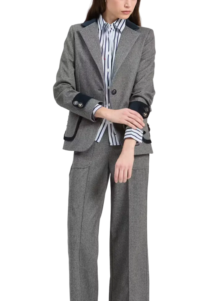 A model wearing a gray herringbone tweed suit with dark navy contrast trim on the jacket and trousers, styled with a blue and white striped shirt and burgundy loafers, against a plain light gray background.