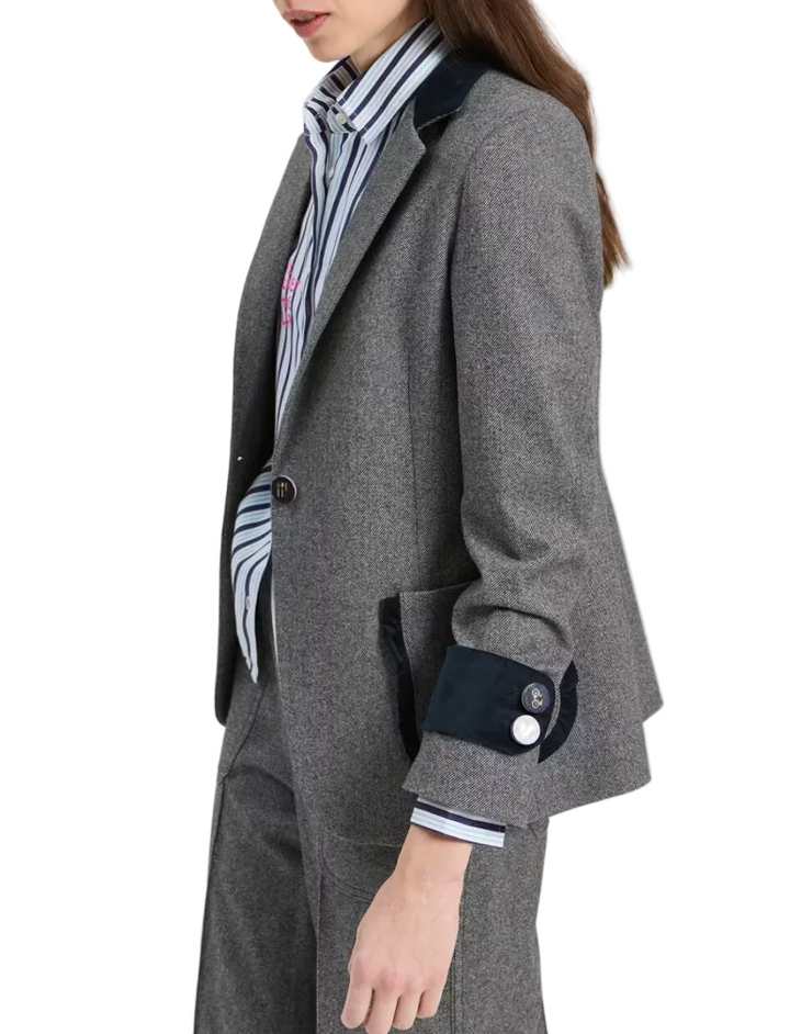 A woman wearing a gray textured blazer with dark navy velvet trim on the collar and cuffs, paired with a striped shirt and matching gray trousers, shown from the chest down against a light gray studio background.
