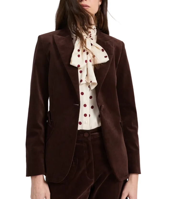 A model wearing a dark brown corduroy blazer and matching trousers, paired with a cream blouse featuring a large bow and red polka dots, against a light gray studio background.
