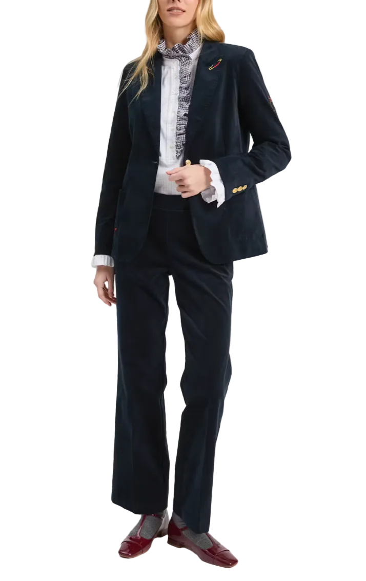 A model wears a dark navy corduroy suit consisting of a blazer and wide-leg trousers, styled with a white patterned high-neck blouse, gold buttons, and dark red patent leather loafers against a light gray studio background.