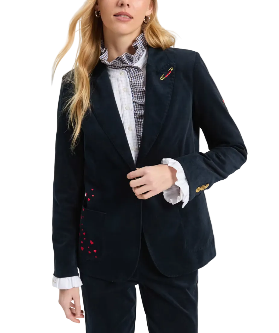 A woman models a dark navy corduroy blazer with red heart embroidery on the pocket, styled with a checkered scarf and white ruffled shirt cuffs, against a light gray studio background.