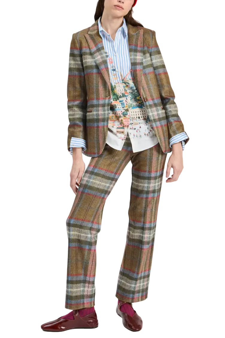 A model wearing a brown plaid blazer and matching trousers, styled with a blue striped shirt, a colorful graphic print vest, and burgundy loafers, against a plain light gray background.