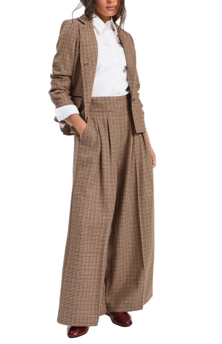 A model wearing a brown and beige houndstooth blazer and matching wide-leg pleated trousers, styled with a white collared shirt and brown leather shoes, against a light gray studio background.