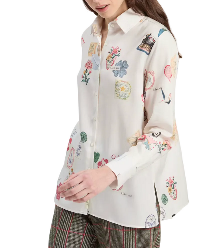 A woman wearing a cream-colored long-sleeve button-up shirt with a colorful whimsical print of hearts, flowers, and celestial motifs, paired with brown plaid trousers against a light gray studio background.