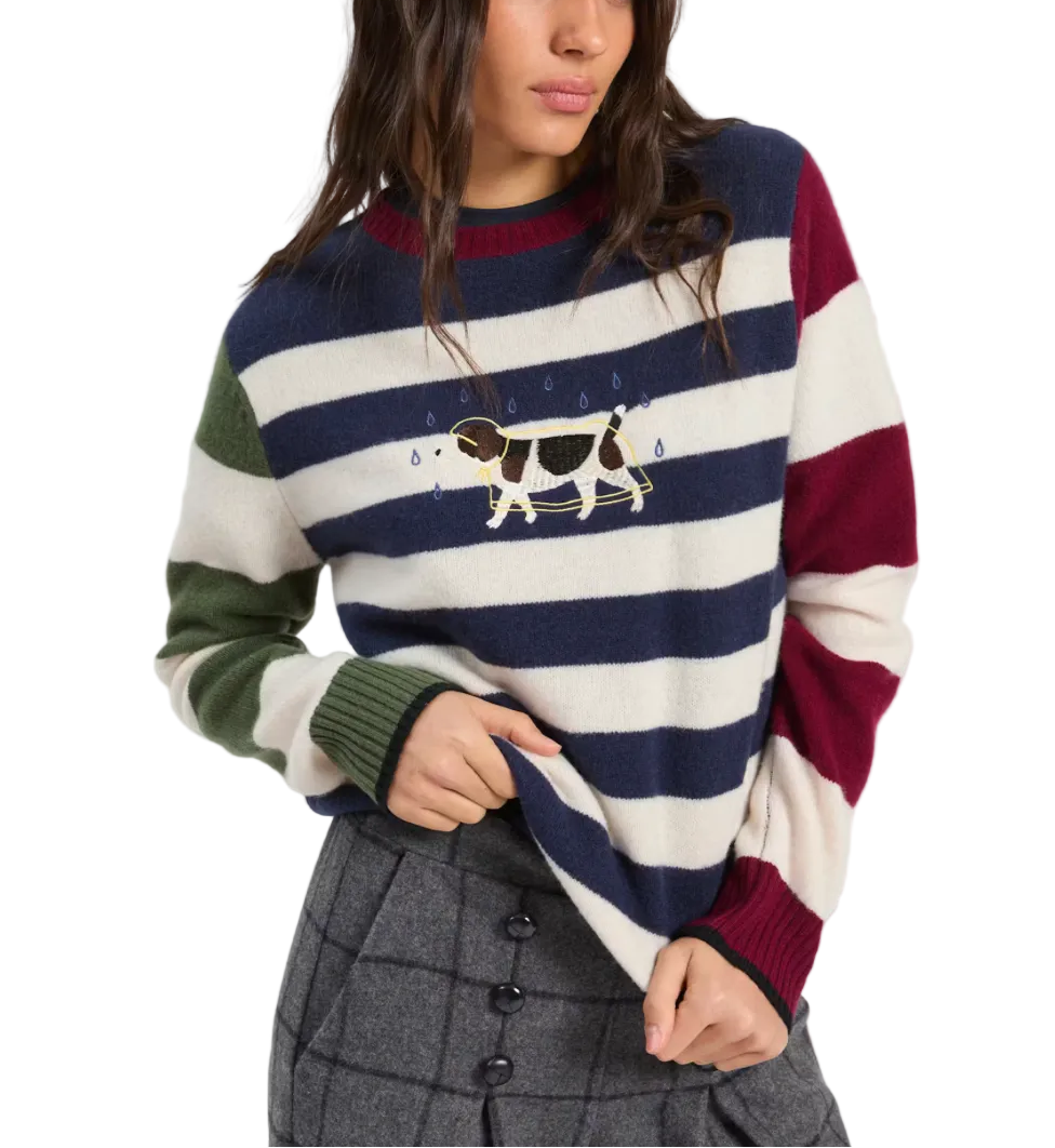 A woman wearing a striped knit sweater with navy, cream, green, and burgundy horizontal bands, featuring a cow graphic with raindrops across the chest. She is paired with a gray plaid skirt against a light gray studio background.