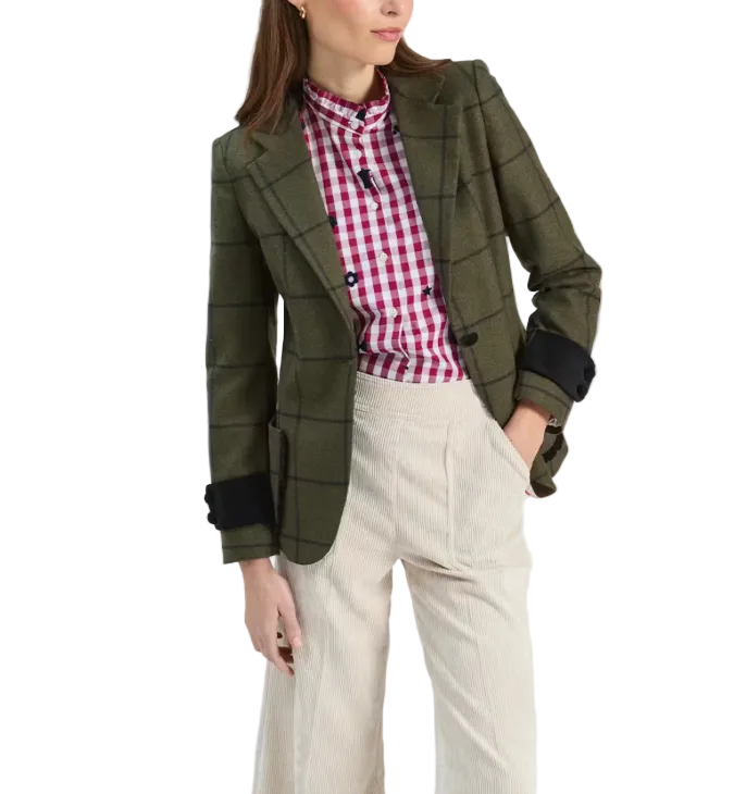 A woman models an olive green windowpane plaid blazer with black cuff tabs, layered over a red and white gingham shirt and paired with cream-colored corduroy trousers, against a plain white studio background.