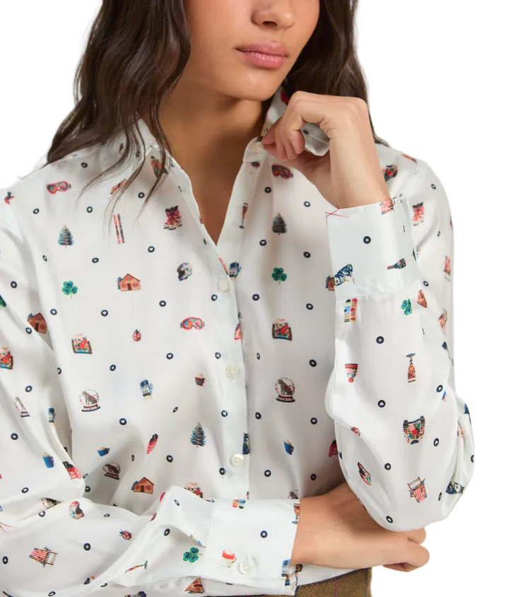 A woman wearing a white button-up blouse with a colorful allover print of small houses, trees, and dots, shown from the chest up against a light gray studio background.