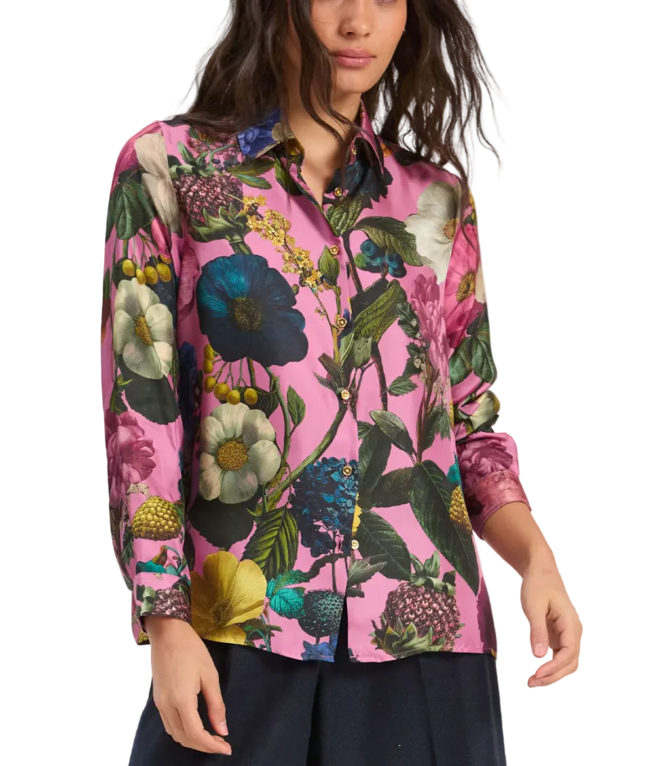 A woman wearing a long-sleeve button-up shirt with a vibrant pink background and a dense multicolor botanical print featuring blue, green, yellow, and white flowers, paired with dark navy pants against a light gray studio background.
