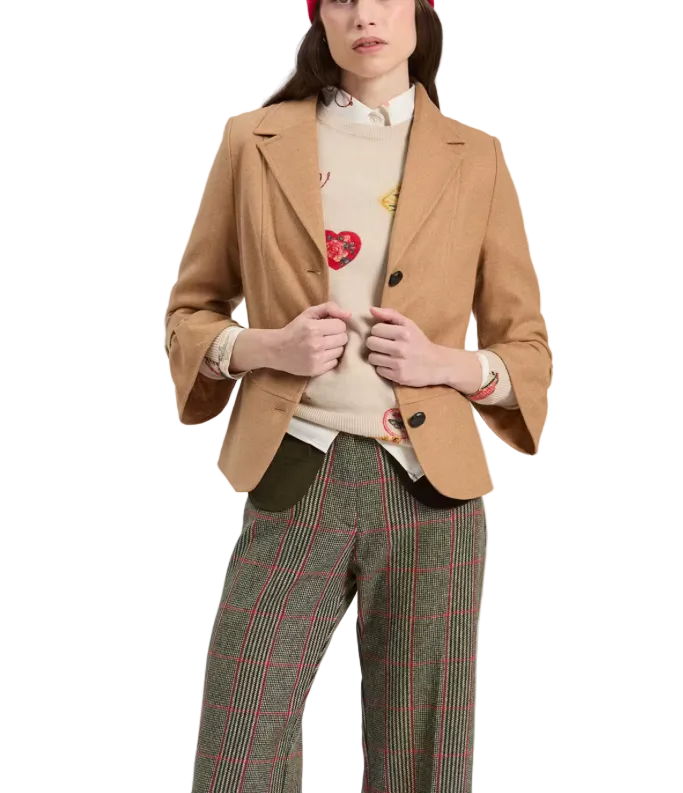 A model wearing a camel wool blazer over a cream knit sweater with colorful heart patches, paired with green plaid trousers and a red beret, against a plain light gray background.