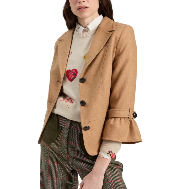 A model wearing a camel brown tailored blazer with a ruffled cuff over a cream knit sweater with a red heart motif, paired with green plaid trousers, against a plain light gray background.