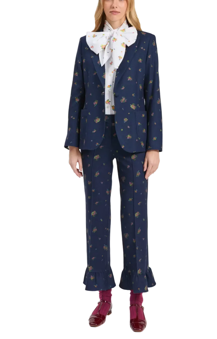A model wearing a navy blue two-piece suit with a small gold floral pattern, styled with a white bow tie, a white shirt, and burgundy shoes against a light gray background.