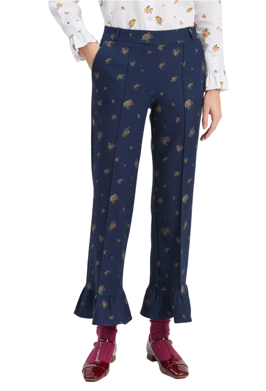 A model is shown from the waist down wearing navy blue trousers with a small multicolor floral print, paired with a white floral blouse, burgundy socks, and burgundy leather shoes against a light gray studio background.