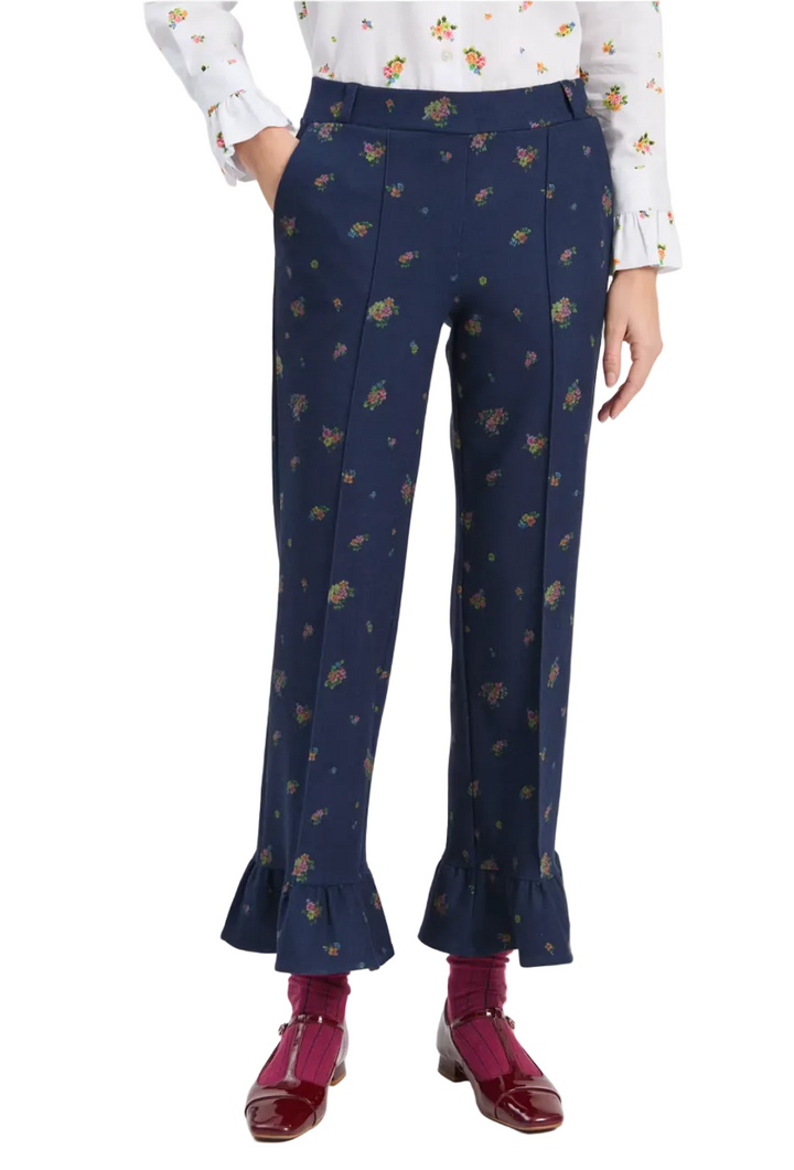 A model is shown from the waist down wearing navy blue trousers with a small multicolor floral print, paired with a white floral blouse, burgundy socks, and burgundy leather shoes against a light gray studio background.