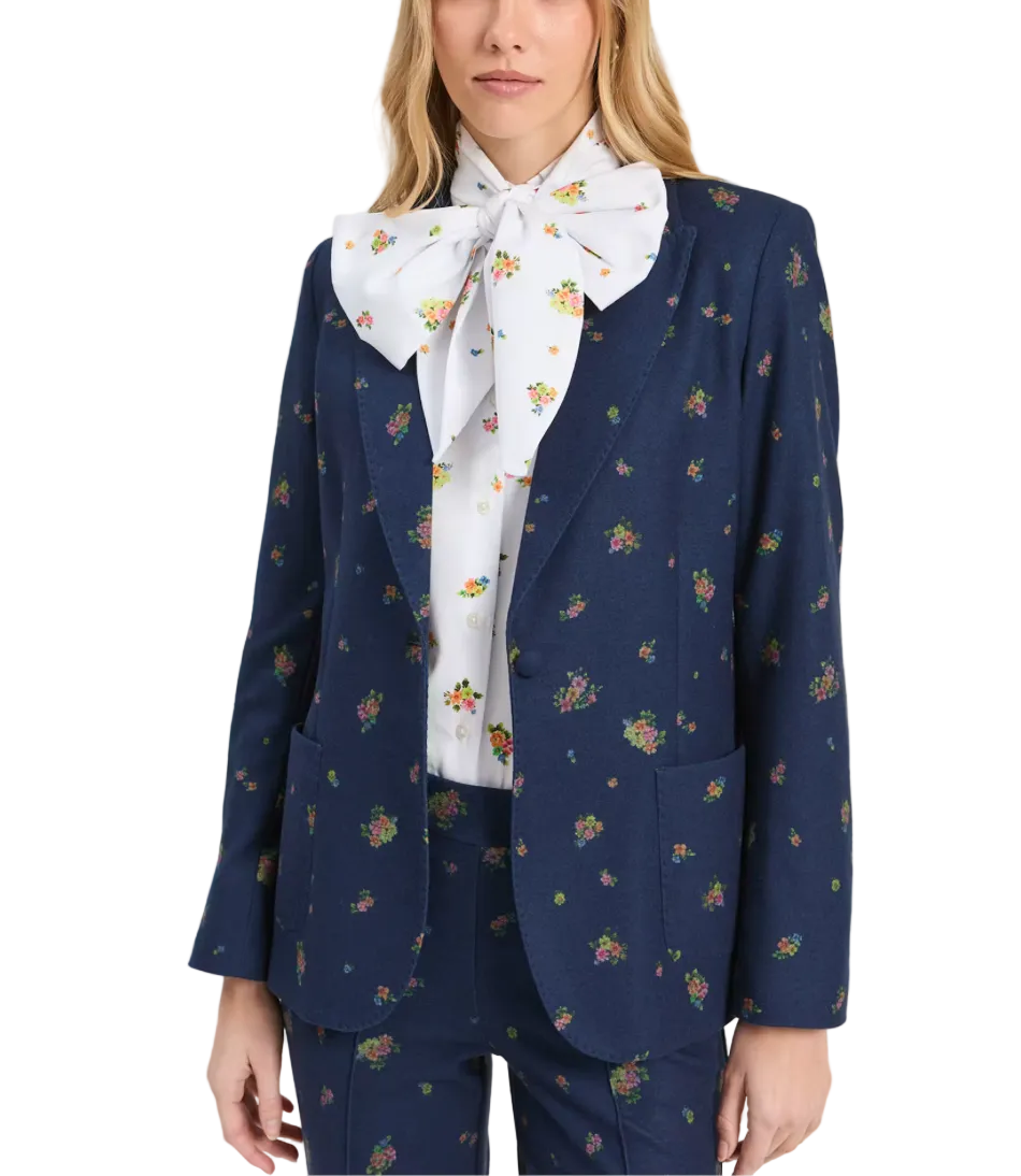 A model wearing a navy blue floral patterned blazer with matching trousers, styled with a white bow tie shirt featuring a small floral print, against a plain light gray background.
