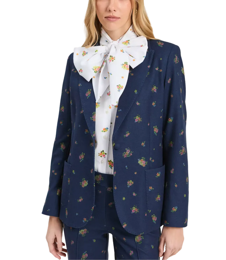A model wearing a navy blue floral patterned blazer with matching trousers, styled with a white bow tie shirt featuring a small floral print, against a plain light gray background.