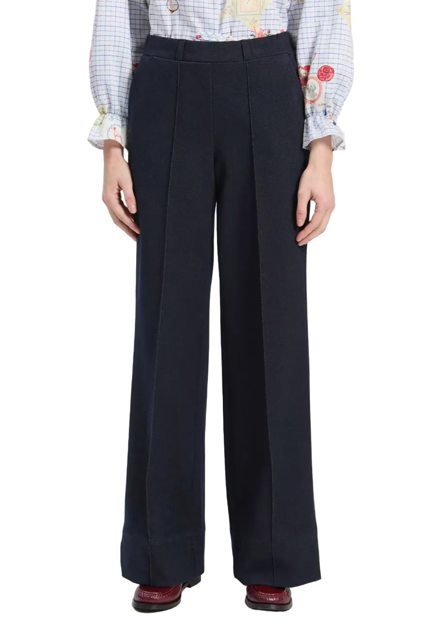 A model is wearing a pair of dark navy blue wide-leg trousers with front pleats, styled with a light blue and white check shirt and burgundy shoes against a plain light gray background.