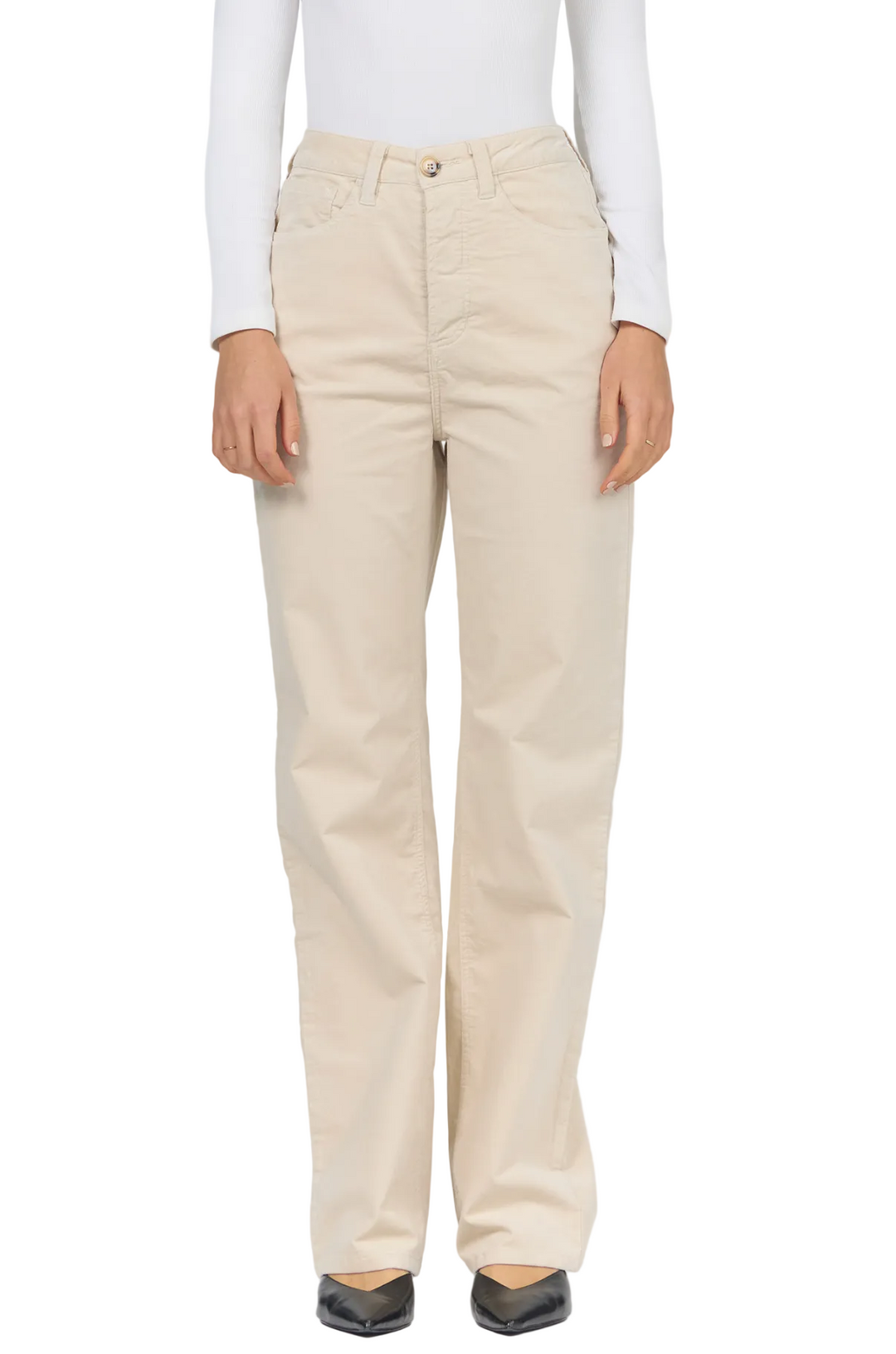 A person wearing straight-leg beige pants with a white long-sleeve top and black pointed flats, shown from the neck down against a light gray studio background.