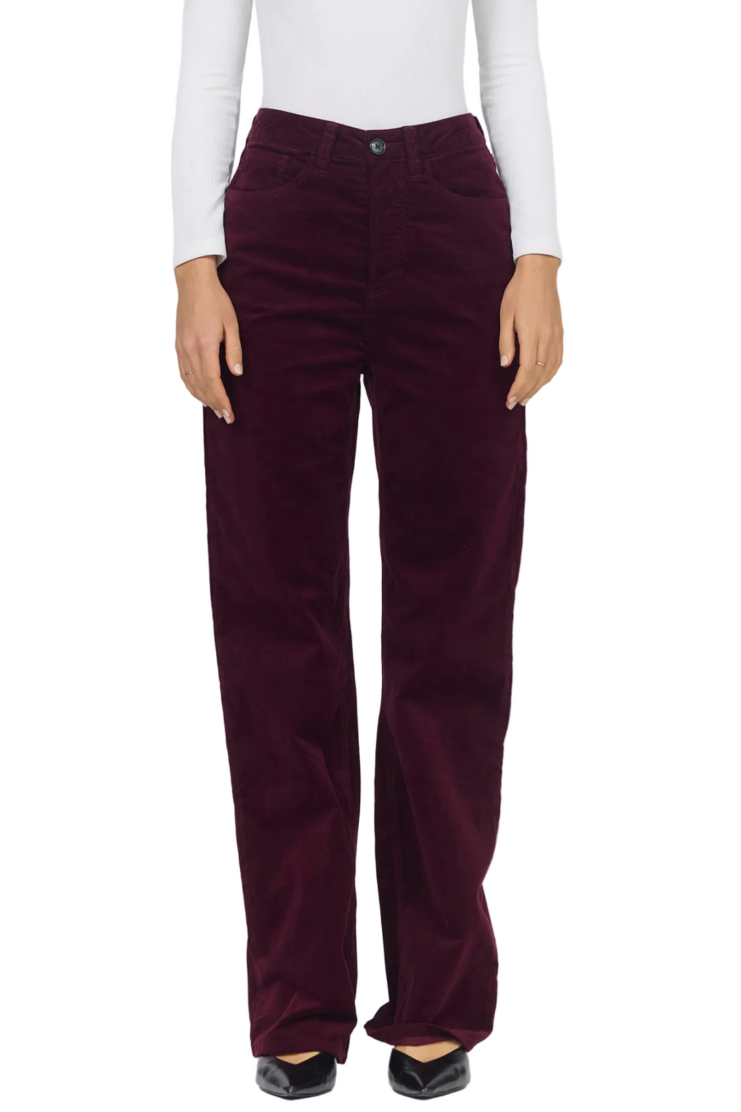 A person wearing dark burgundy corduroy pants with a white long-sleeve top and black pointed flats, shown from the waist down against a light gray studio background.