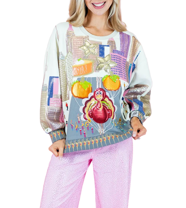 A woman wearing a colorful, patterned crewneck sweatshirt and pink patterned pants, standing against a solid pink studio background.