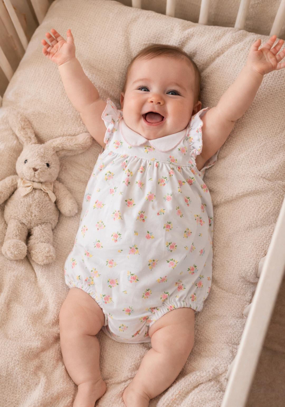 A smiling baby lying on their back in a white romper with a small pink and yellow floral print, surrounded by a beige blanket and a plush bunny toy.