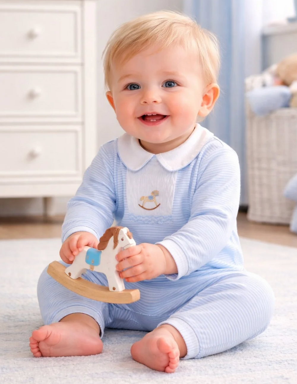 A smiling baby sitting on a light blue rug in a nursery, wearing a light blue and white striped long-sleeve romper with a white Peter Pan collar. The baby is holding a wooden rocking horse toy.