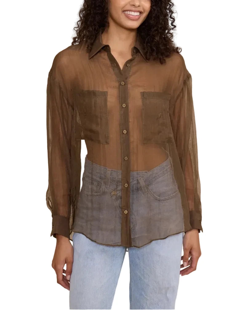 A woman modeling a sheer brown button-up shirt with a layered, asymmetrical design over a gray denim-style vest, paired with light blue jeans, against a solid beige background.