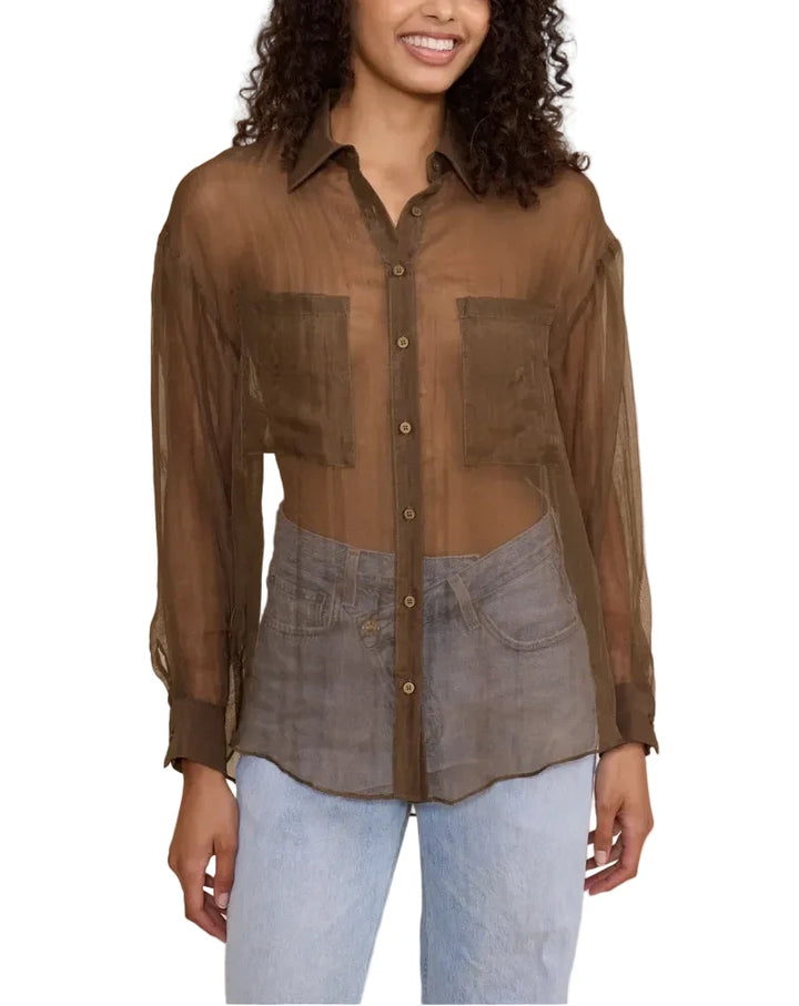A woman modeling a sheer brown button-up shirt with a layered, asymmetrical design over a gray denim-style vest, paired with light blue jeans, against a solid beige background.