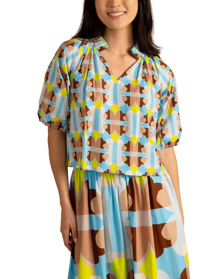 Woman wearing the Trish Top in Small Deco by Double A, a colorful patterned top on a beige background