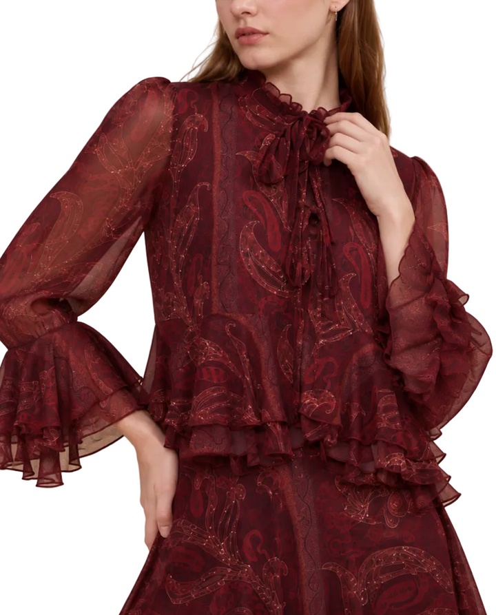 A woman wearing a sheer burgundy long-sleeve blouse with a paisley pattern, ruffled collar, and tie detail, paired with a matching skirt, set against a solid beige background.