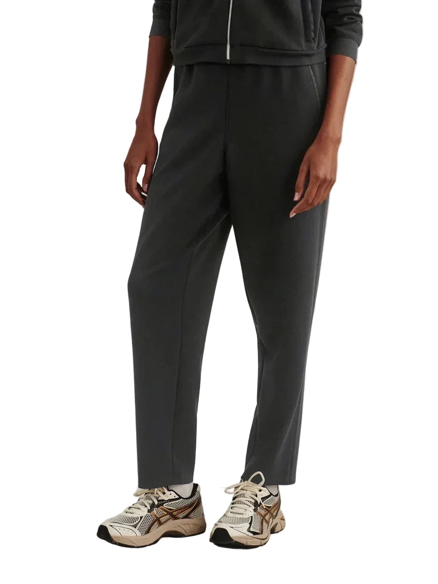 A model wearing dark charcoal gray jogger pants with a matching zip-up hoodie and white and beige athletic sneakers, standing against a neutral beige studio background.