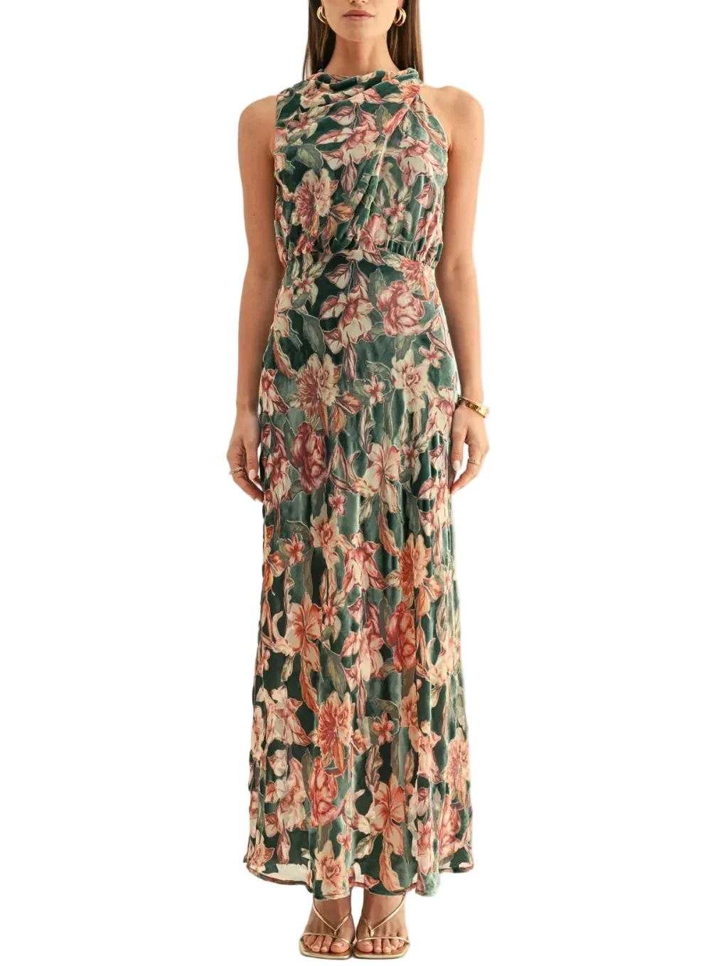 A woman modeling a sleeveless halter-neck maxi dress with a green base and a pink and peach floral pattern, set against a plain off-white studio background.