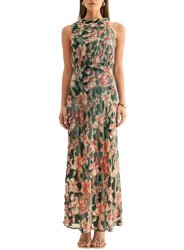 A woman modeling a sleeveless halter-neck maxi dress with a green base and a pink and peach floral pattern, set against a plain off-white studio background.