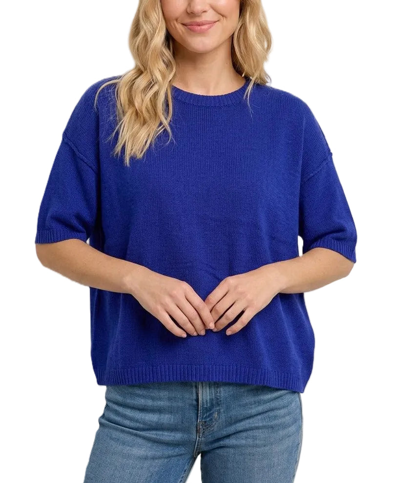 A woman wearing a vibrant royal blue short-sleeve knit sweater and blue jeans, standing against a solid light beige background.