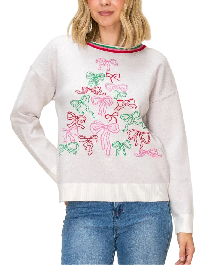 A woman wearing a white crewneck sweatshirt decorated with a colorful embroidered Christmas tree made of pink and green bows, paired with blue jeans, against a solid beige background.
