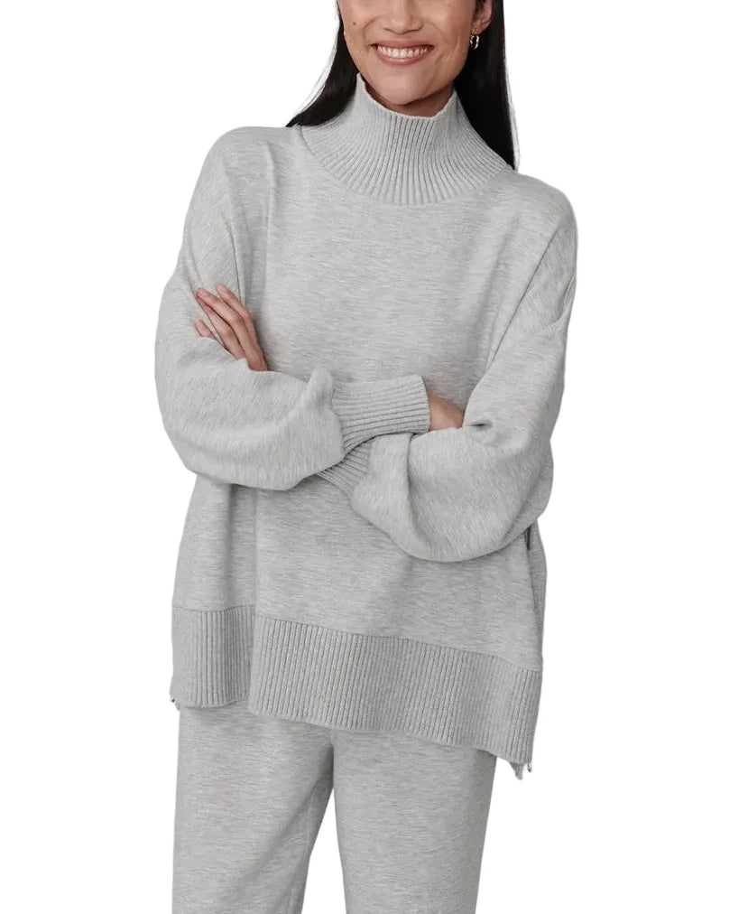 A woman modeling a light gray heathered knit turtleneck sweater and matching sweatpants against a plain light gray studio background.
