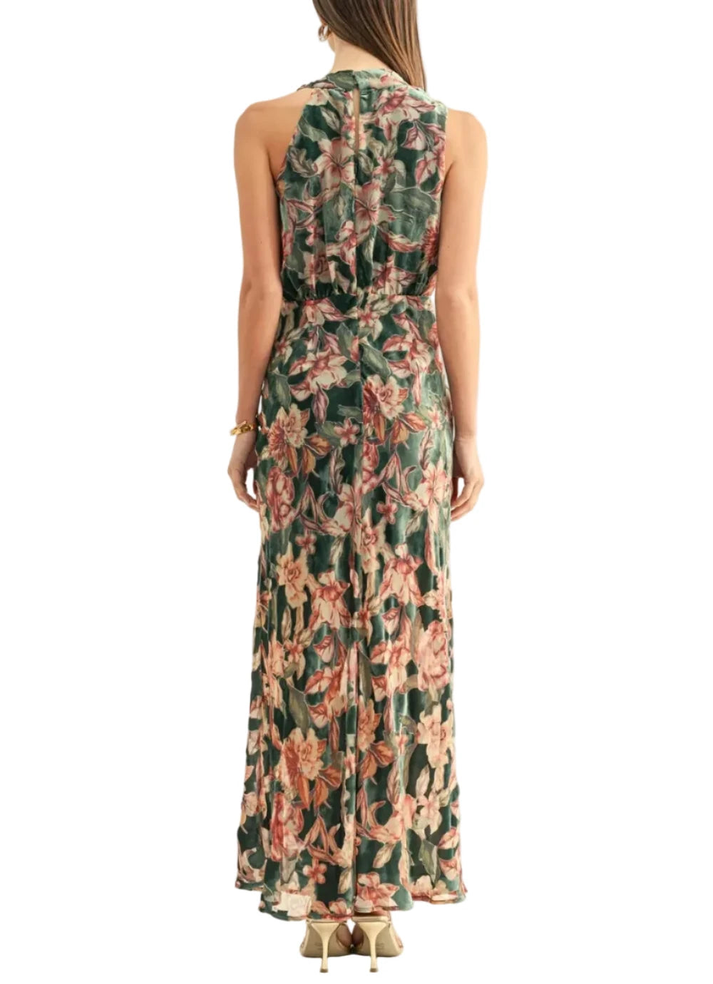A woman viewed from the back wearing a sleeveless maxi dress with a tropical floral print in green, pink, and beige tones, paired with beige heels against a plain white studio background.