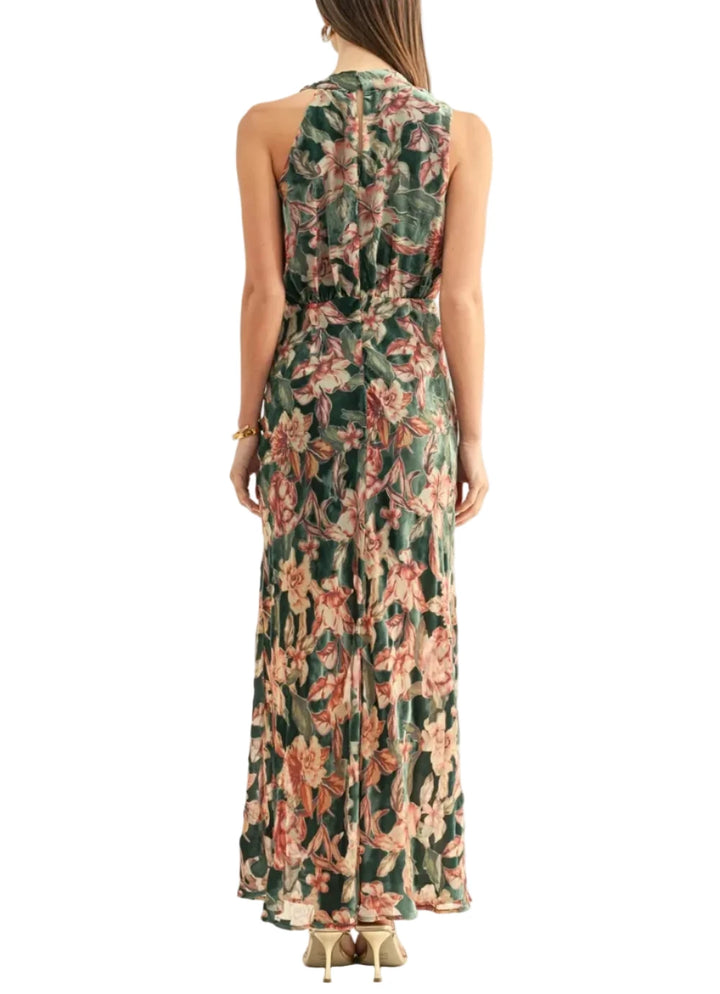 A woman viewed from the back wearing a sleeveless maxi dress with a tropical floral print in green, pink, and beige tones, paired with beige heels against a plain white studio background.