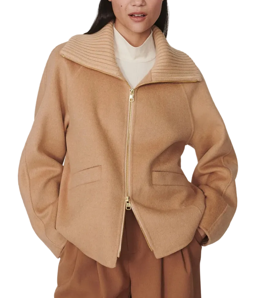 A woman modeling a camel-colored zip-up jacket with a wide ribbed collar, worn over a cream turtleneck and brown trousers, against a plain light gray studio background.