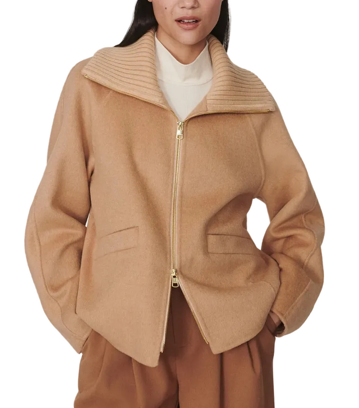 A woman modeling a camel-colored zip-up jacket with a wide ribbed collar, worn over a cream turtleneck and brown trousers, against a plain light gray studio background.