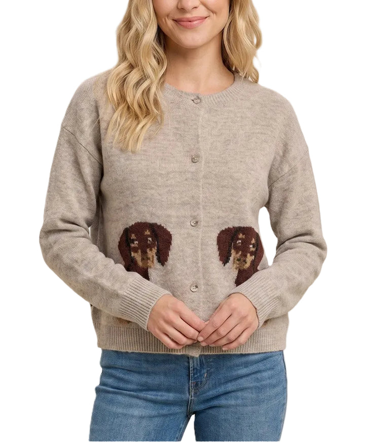A woman wearing a light beige knit cardigan sweater with two brown dog face patches on the lower front, paired with blue jeans, against a solid light beige background.