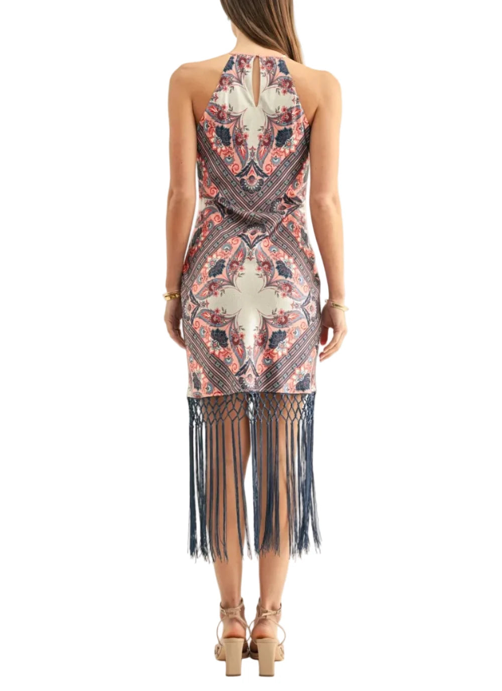 A woman viewed from the back wearing a sleeveless halter-neck dress with a navy, coral, and beige bohemian-style pattern and a long fringe hem, paired with tan strappy heels against a plain white background.