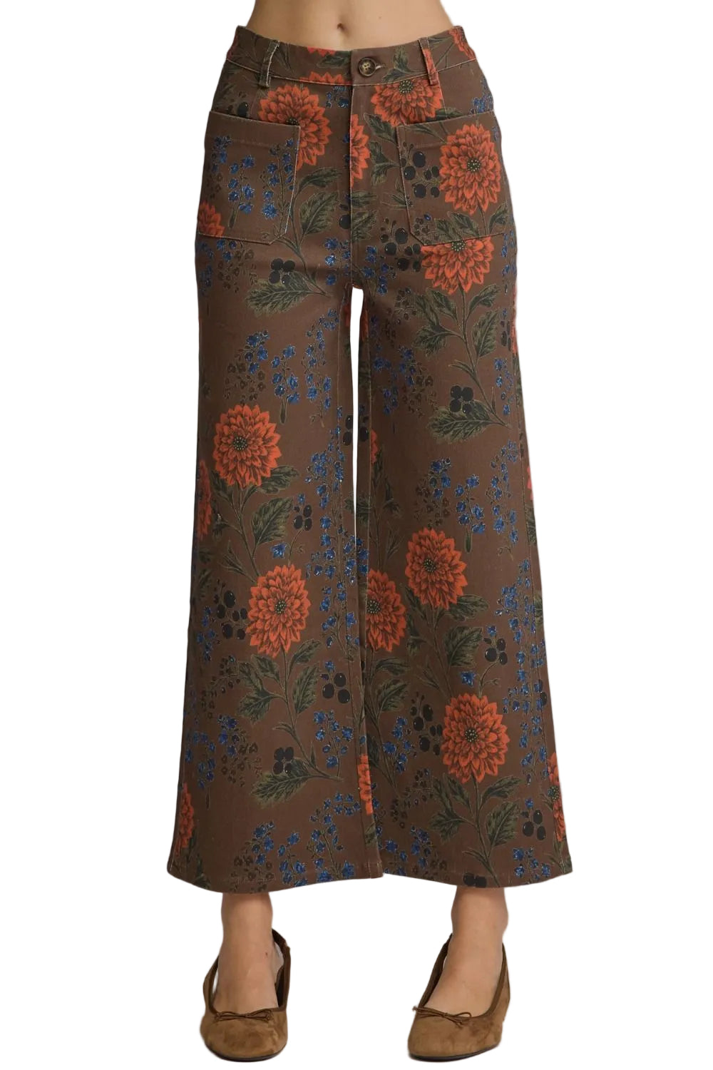 A model is wearing a pair of brown wide-leg cropped pants with a vibrant orange, blue, and green floral pattern, paired with brown suede flats, shot against a solid gray studio background.