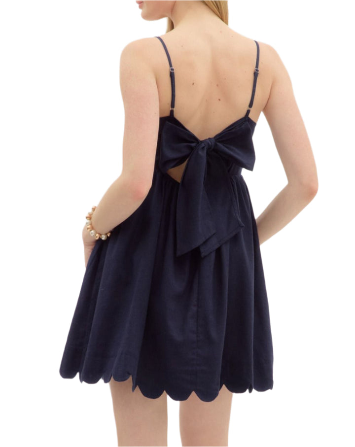 Solid adjustable shoulder strap pullover mini dress featuring scallop accent neck and hem finish. Elasticated skirt waist and bow tie closure at back. Pockets at sides. Lined. Woven. Non-sheer. Lightweight.