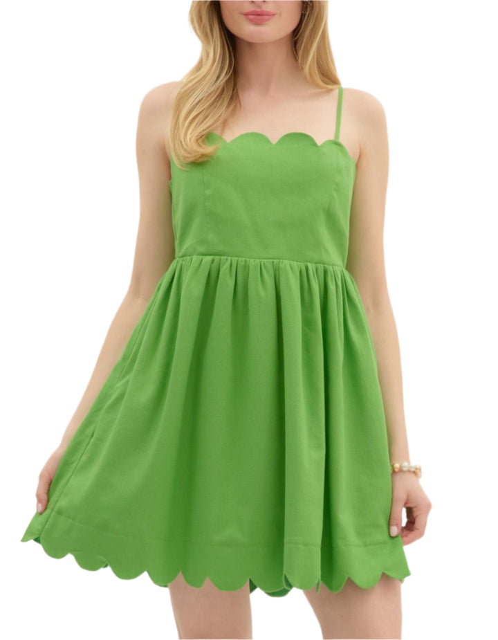 Solid adjustable shoulder strap pullover mini dress featuring scallop accent neck and hem finish. Elasticated skirt waist and bow tie closure at back. Pockets at sides. Lined. Woven. Non-sheer. Lightweight.