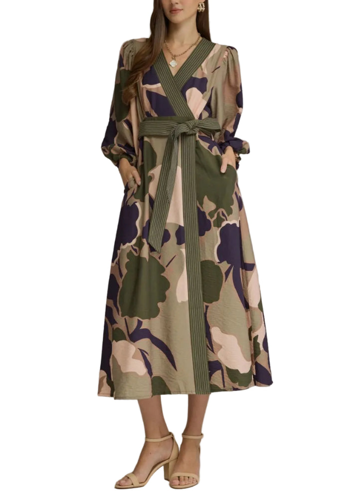 A woman models a long-sleeved midi wrap dress with a large abstract floral print in olive green, beige, and navy blue, featuring a matching waist tie belt, set against a solid gray studio background.