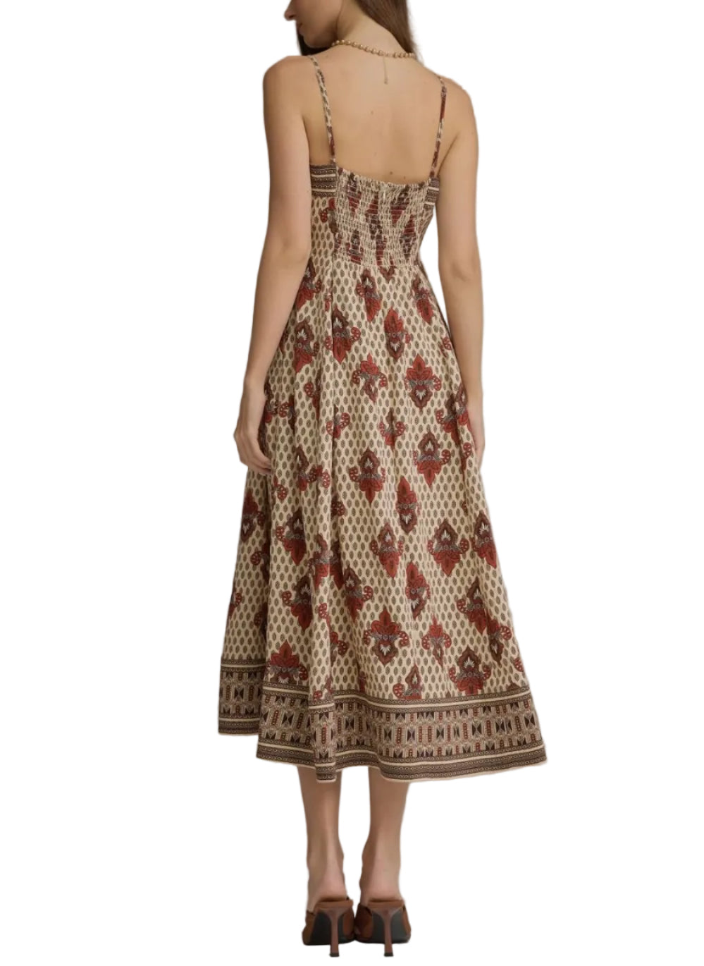 A woman viewed from the back wearing a sleeveless midi dress with thin straps, a smocked bodice, and an allover brown and beige geometric print. She is standing against a plain gray studio background.