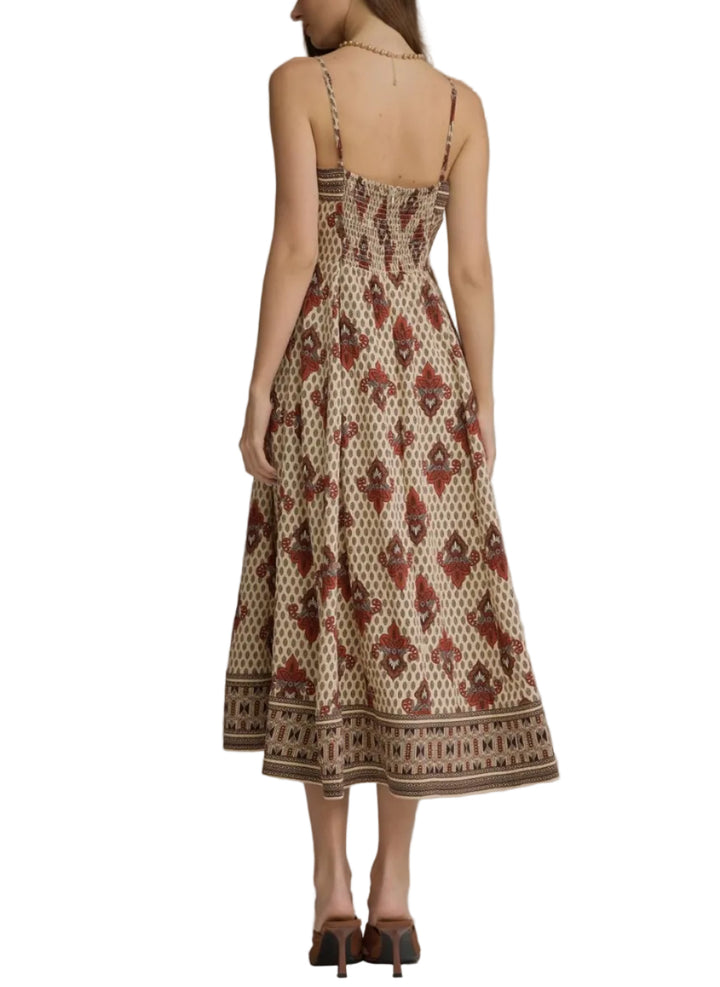 A woman viewed from the back wearing a sleeveless midi dress with thin straps, a smocked bodice, and an allover brown and beige geometric print. She is standing against a plain gray studio background.